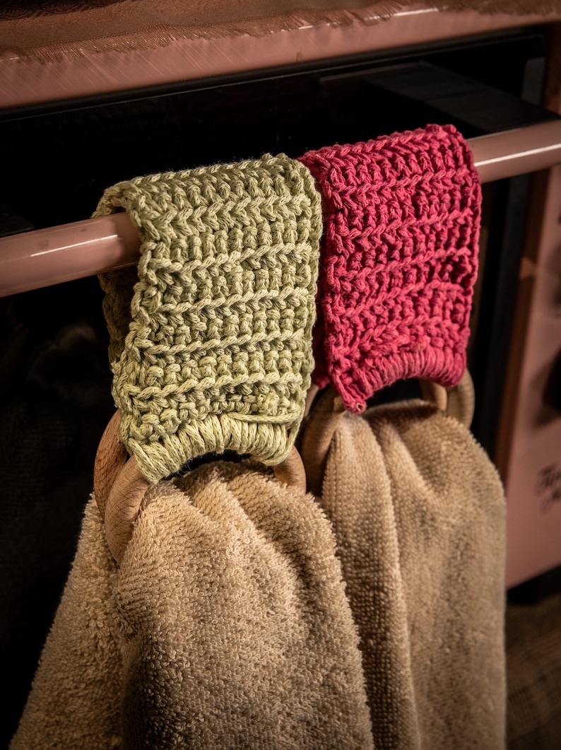 Puede incluir: Dos pa&ntilde;os de cocina colgados de una barra. Las toallas son beige y tienen aros de madera. Los soportes est&aacute;n tejidos a ganchillo, uno en color verde claro y otro en color rosa brillante.
