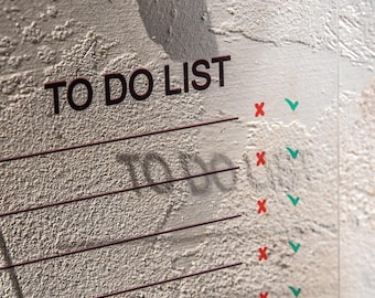 May include: A clear acrylic To Do List with the words "TO DO LIST" in dark brown. The list has horizontal lines for writing tasks, with red X's and green check marks. The background is a textured, off-white wall.
