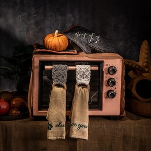 Puede incluir: Un horno rosa de encimera con una peque&ntilde;a calabaza naranja, un guante de horno y dos toallas colgantes. Las toallas tienen la parte superior decorativa y aros de madera. Una toalla dice "olive you".