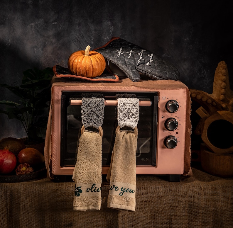 Puede incluir: Un horno rosa de encimera con una peque&ntilde;a calabaza naranja, un guante de horno y dos toallas colgantes. Las toallas tienen la parte superior decorativa y aros de madera. Una toalla dice "olive you".