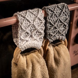 Könnte beinhalten: Zwei Küchentücher hängen an einem rosa Ofengriff. Die beigen Handtücher werden von Holzringen gehalten. Die Halter sind gehäkelt, einer hellgrau und der andere dunkelgrau, mit einem Rautenmuster.