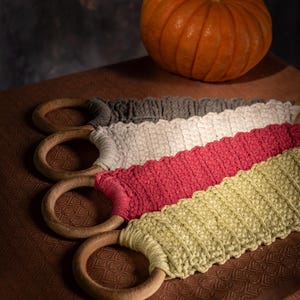 Puede incluir: Cuatro coloridas envolturas de asa de ganchillo con anillos de madera, en gris, blanco, rosa y verde. Una peque&ntilde;a calabaza naranja se encuentra en el fondo. Las envolturas son para bolsas o cestas.
