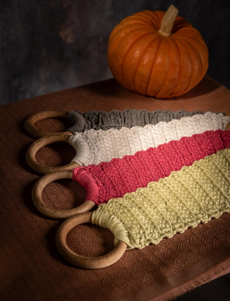 Puede incluir: Cuatro coloridas envolturas de asa de ganchillo con anillos de madera, en gris, blanco, rosa y verde. Una peque&ntilde;a calabaza naranja se encuentra en el fondo. Las envolturas son para bolsas o cestas.