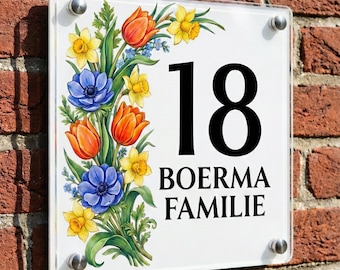 Puede incluir: Un letrero cuadrado para número de casa con el número 18 y el texto BOERMA FAMILIE en fuente negra. El letrero es de acrílico transparente con cuatro soportes metálicos plateados y muestra un colorido arreglo floral de tulipanes y narcisos.
