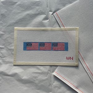 Puede incluir: Un lienzo rectangular de punto de aguja con tres banderas americanas en rojo, blanco y azul. Las banderas están cosidas sobre un fondo gris claro con un borde azul. Las iniciales "WN" están cosidas en rojo en la esquina inferior derecha.