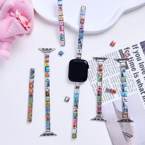 Puede incluir: Un reloj inteligente negro con correas decorativas con pequeños dijes cuadrados con varios diseños. Varias correas y dijes adicionales están dispersos alrededor del reloj.