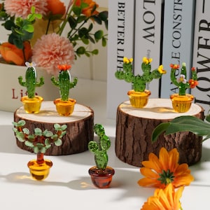 Puede incluir: Figuras de cactus de vidrio en varios estilos, cada una en un recipiente amarillo o marrón. Los cactus tienen cuerpos verdes con detalles florales coloridos. Se exhiben sobre rodajas de madera, con flores en el fondo.
