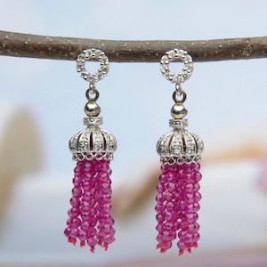 May include: A pair of silver tassel earrings. Each earring has a crown-shaped top with clear gemstones, a silver ball, and a cascade of small, magenta, faceted beads. The earrings are displayed on a thin, brown branch against a soft, blurred background.