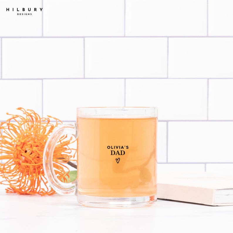 May include: A clear glass mug filled with a light orange liquid, featuring the text "OLIVIA'S DAD" with a heart symbol. An orange flower and a white notebook are also in the image. The mug has a handle and is set against a white tile background.