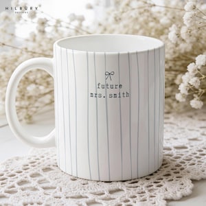May include: A white ceramic mug featuring thin vertical light blue stripes. The front of the mug displays a small black line-art bow icon above the black typewriter-style text that reads future mrs. smith. The mug has a curved handle.