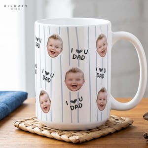 May include: A white ceramic mug featuring a repeating pattern of light blue vertical stripes, circular photographs of a smiling baby, and the black text I heart U DAD. The mug has a large handle on the right side and a smooth, glossy finish.