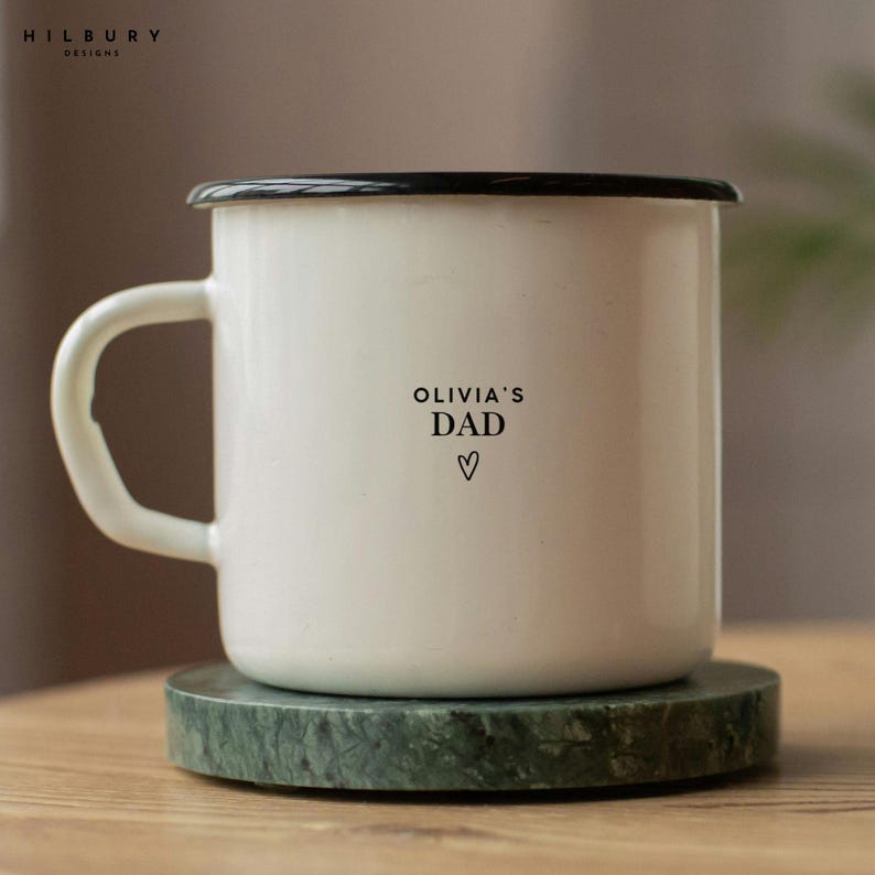 May include: A white enamel mug with a black rim and handle, resting on a green marble coaster. The mug features the text "OLIVIA'S DAD" with a heart symbol below. The mug is a perfect gift for a father.