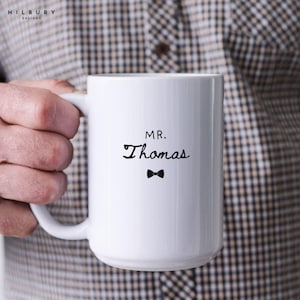 Może przedstawiać: Biały ceramiczny kubek z napisem "MR. Thomas" i małą czarną muchą. Kubek ma duży uchwyt i prosty, elegancki design, idealny na spersonalizowany prezent.