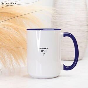 May include: White ceramic mug with a navy blue rim and handle. The mug features the text "Olivia's Dad" with a heart symbol. The mug is a standard size for coffee or tea.