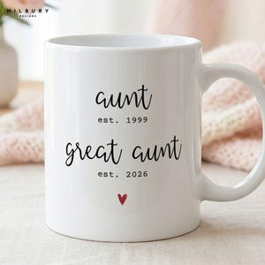 May include: A white ceramic mug featuring black text that reads aunt est. 1999 and great aunt est. 2026, with a small red heart icon centered below the text. The mug has a smooth finish and a curved handle on the right side for holding.
