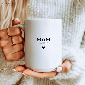 Könnte beinhalten: Weißer Keramikbecher mit dem Wort „MOM“, dem Jahr „est. 1990“ und einem schwarzen Herz. Der Becher wird von einer Person gehalten. Der Becher hat einen großen Henkel und eine zylindrische Form. Der Becher ist ein Geschenk für eine Mutter.