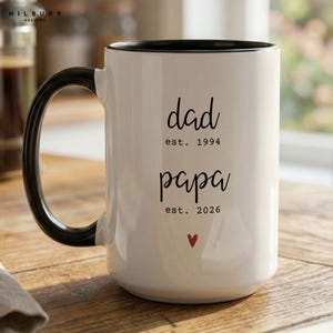 Peut inclure: Une tasse en céramique blanche avec une anse et un bord noirs. La tasse porte les mots "dad est. 1994" et "papa est. 2026" en écriture noire, avec un petit cœur rouge. La tasse est posée sur une surface en bois.