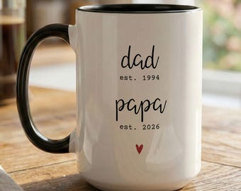 May include: A white ceramic mug with a black handle and rim. The mug features the words "dad est. 1994" and "papa est. 2026" in black script, with a small red heart. The mug is on a wooden surface.