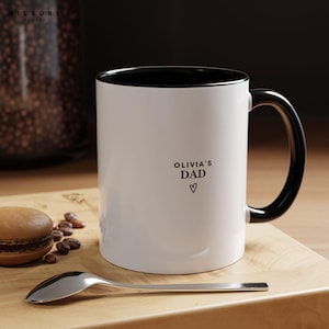 May include: White ceramic mug with a black interior and handle. The mug has the text "OLIVIA'S DAD" with a heart symbol. A silver spoon and a macaron are on a wooden surface next to the mug.