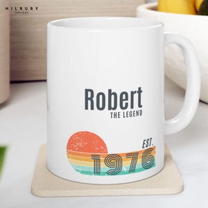 May include: White ceramic mug with the name "Robert" and the text "THE LEGEND" in dark gray. Below is a retro design of a sun with the year "1976" in a vintage font. The mug is on a beige coaster.
