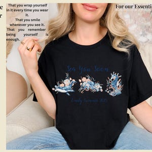 May include: A black short-sleeved t-shirt featuring a graphic print of a whale, an octopus, and a seahorse surrounded by floral elements. The text reads Sea You Soon and Emily Summer 2025. The shirt has a crew neckline and a standard fit design.