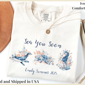 May include: An ivory Comfort Colors t-shirt featuring a watercolor graphic of a whale, octopus, and seahorse surrounded by coral. The text reads Sea You Soon and Emily Summer 2025 in blue script. A gold necklace rests on the neckline of the shirt.