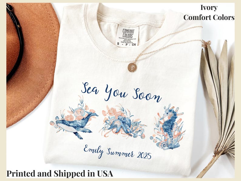 May include: An ivory Comfort Colors t-shirt featuring a watercolor graphic of a whale, octopus, and seahorse surrounded by coral. The text reads Sea You Soon and Emily Summer 2025 in blue script. A gold necklace rests on the neckline of the shirt.