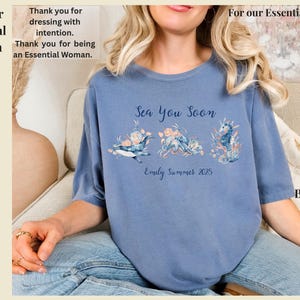 May include: A blue jean colored t-shirt featuring a graphic print with the text Sea You Soon and Emily Summer 2025. The design includes illustrations of a whale, an octopus, and a seahorse surrounded by floral elements in shades of blue and peach.
