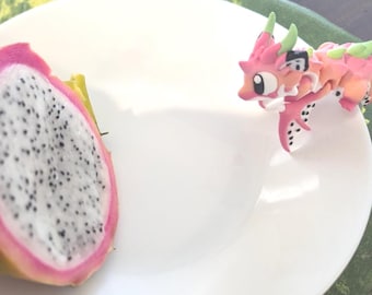 May include: A halved dragon fruit with a pink rind and white flesh speckled with black seeds sits on a white plate. A small, pink and orange dragon figurine with green accents is also on the plate.