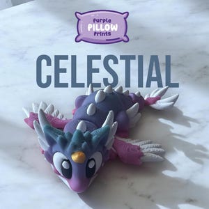 Puede incluir: Una figurita de dragón impresa en 3D de color morado, azul y blanco con un cuerno dorado. El dragón está acostado boca abajo con las alas extendidas. La figurita está etiquetada como "Celestial" y "Purple Pillow Prints".