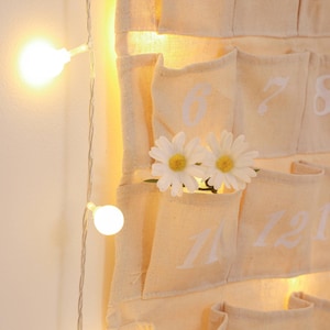 May include: A beige canvas advent calendar with white numbers 6, 7, 8, 11, and 12. The calendar is decorated with two white daisies and a string of white lights.