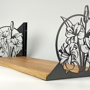 Puede incluir: Estante de madera con soportes metálicos negros con un diseño floral. Los soportes tienen un diseño circular con flores de lirio recortadas. El estante es de madera marrón claro y está montado sobre un fondo blanco.