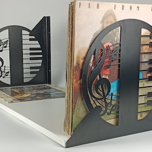 May include: Black metal record holder with a musical note and piano key design. The holder is on a white shelf and holds vinyl records. The design includes a treble clef, musical notes, and piano keys.