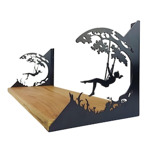 Op de afbeelding: Een decoratief wandrek met een houten plank en zwarte metalen steunen. De steunen hebben een silhouet van een persoon op een schommel onder een boom. Het rek is ontworpen om aan de muur te worden bevestigd.