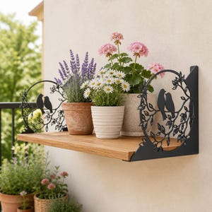 Könnte beinhalten: Ein Wandregal aus Holz mit schwarzen Metallhalterungen, die ein kreisförmiges Design von Vögeln auf Zweigen zeigen. Auf dem Regal stehen drei Topfpflanzen: eine mit violettem Lavendel, eine mit weißen Gänseblümchen und eine mit rosa Geranien.
