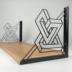 May include: A wooden shelf with black metal brackets featuring an optical illusion design. The shelf is made of light-colored wood and is supported by two black metal brackets with geometric shapes. The brackets are mounted on a white wall.