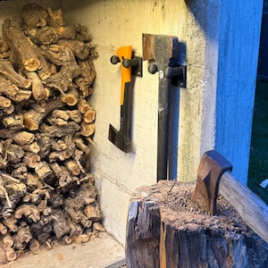 Puede incluir: Una escena al aire libre con una pila de leña y dos hachas montadas en una pared de hormigón. Un hacha tiene un mango naranja, la otra un mango negro. Un hacha está incrustada en un tocón de árbol en primer plano.