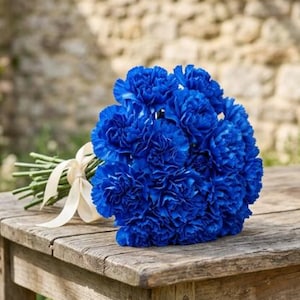 May include: A bouquet of vibrant blue carnations tied with a cream-colored ribbon. The flowers are arranged on a rustic wooden table, creating a striking contrast of colors and textures. The background is blurred, suggesting an outdoor setting.