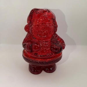 May include: A translucent red glass figurine shaped like Santa Claus. The figure features molded details including a hat, a full beard, a coat with a textured belt, and boots. The glass has a glossy finish and captures light, creating deep red tones.