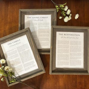 Puede incluir: Tres impresiones enmarcadas con texto. Los marcos son de madera gris desgastada. El texto de las impresiones incluye "The Living Christ", "The Family" y "The Restoration". Flores blancas en primer plano.