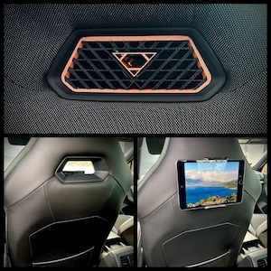 Op de afbeelding: Auto-interieur met een koperkleurige geometrische ventilatie en een zwart ruitpatroon. Daaronder een zwarte leren autostoel met een tablet op de hoofdsteun, met een landschap.