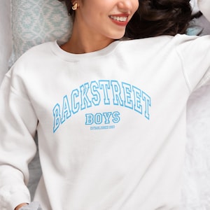 Könnte beinhalten: Weißes Sweatshirt mit Rundhalsausschnitt und dem Text "BACKSTREET BOYS" in Hellblau, der sich über die Brust wölbt. Unter dem Bandnamen stehen "BOYS" und "ESTABLISHED 1993" in kleinerer Schrift. Lässiger Stil.