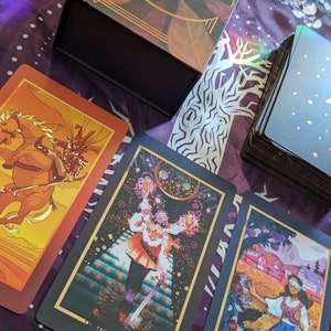 Może przedstawiać: Trzy karty tarota z talii The Chromatic Fates Tarot. Karty to Mag, Głupiec i Rydwan. Karty leżą na fioletowo-białym wzorzystym materiale.