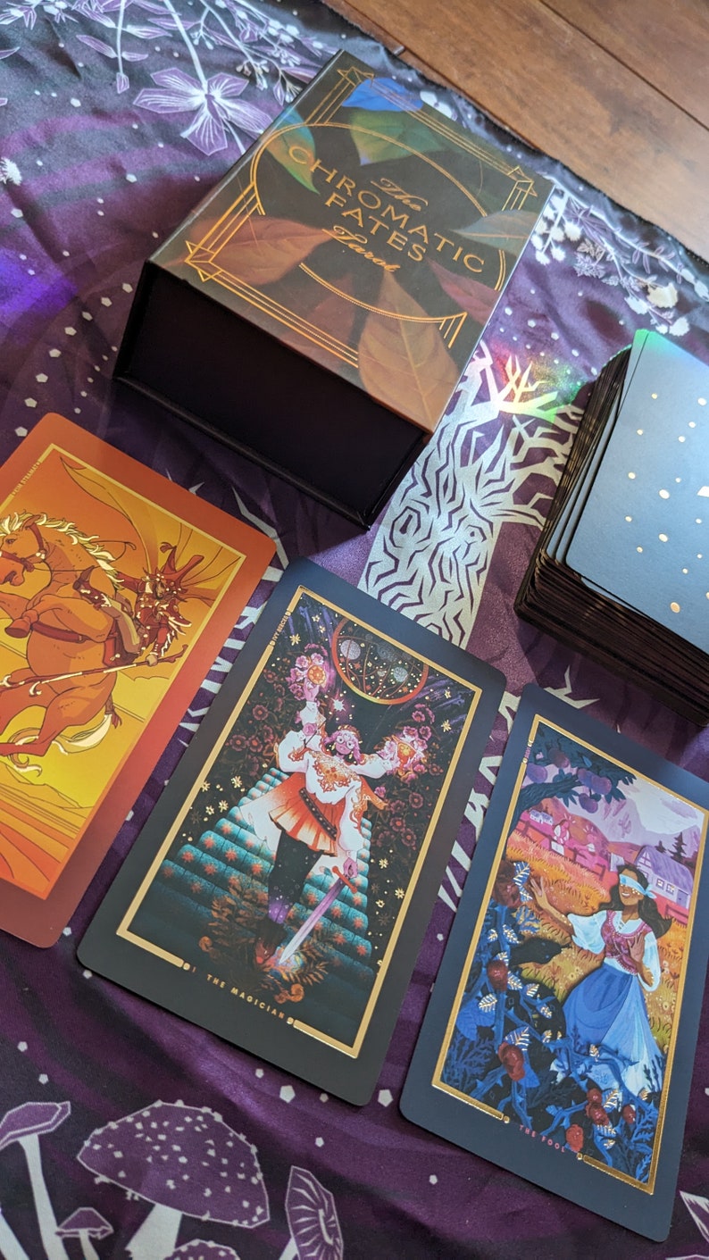 Może przedstawiać: Trzy karty tarota z talii The Chromatic Fates Tarot. Karty to Mag, Głupiec i Rydwan. Karty leżą na fioletowo-białym wzorzystym materiale.