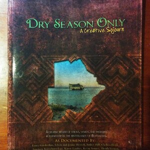 May include: A book cover with a brown background and a torn section revealing a blue sky and a yellow school bus. The title of the book is "Dry Season Only: A Creative Sojourn".