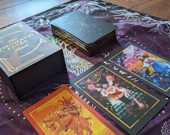 Könnte beinhalten: Ein Deck The Chromatic Fates Tarotkarten in einer schwarzen Schachtel mit goldenen Schriftzügen. Die Karten liegen auf einem lila-weißen gemusterten Stoff ausgebreitet. Die Karten zeigen farbenfrohe Illustrationen und goldene Akzente.