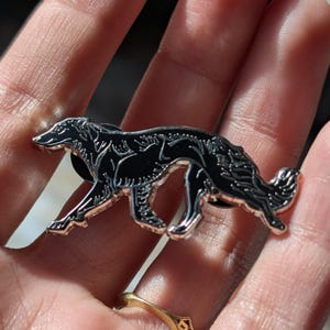 K&ouml;nnte beinhalten: Ein schwarzer und silberner Ansteckpin in Form eines Hundes, wahrscheinlich ein Borzoi, mit aufwendigen Details. Der Pin wird vor einer Hand gezeigt, mit einem goldenen Ring im Hintergrund. Der Hund ist in einer Laufhaltung dargestellt.