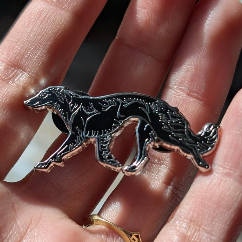 K&ouml;nnte beinhalten: Ein schwarzer und silberner Ansteckpin in Form eines Hundes, wahrscheinlich ein Borzoi, mit aufwendigen Details. Der Pin wird vor einer Hand gezeigt, mit einem goldenen Ring im Hintergrund. Der Hund ist in einer Laufhaltung dargestellt.