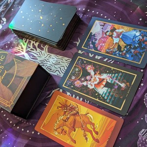 Może przedstawiać: Czarne pudełko z złotymi akcentami i tekstem "The Chromatic Fates Tarot" na przodzie. Pudełko jest otwarte i zawiera talię kart tarota z kolorowymi ilustracjami. Karty są rozłożone na fioletowym materiale z białym wzorem grzyb&oacute;w.