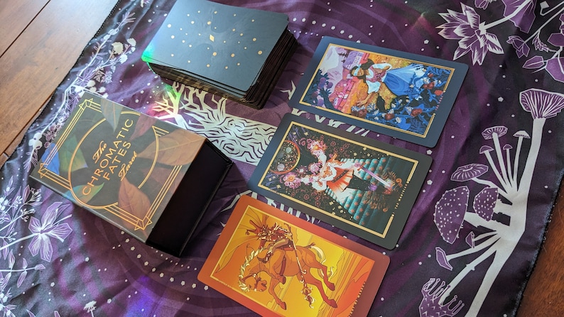 Może przedstawiać: Czarne pudełko z złotymi akcentami i tekstem "The Chromatic Fates Tarot" na przodzie. Pudełko jest otwarte i zawiera talię kart tarota z kolorowymi ilustracjami. Karty są rozłożone na fioletowym materiale z białym wzorem grzyb&oacute;w.